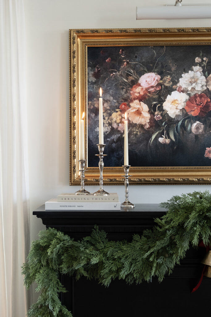 Closeup image of three silver candlesticks with ivory taper candles styled on a black fireplace mantle. The mantle has a cedar garland hung on it and a floral painting above it.
